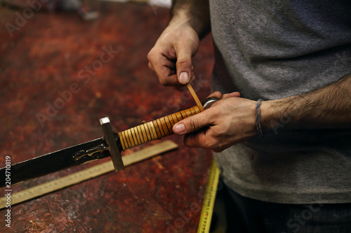 Smith makes leather winding of sword. Man is working in workshop. He fixes leather band on hilt of sword.