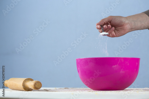 kneading dough in a bright pizza dish. female hand close-up adds sugar with a teaspoon and a wooden rolling pin next to it on a table sprinkled with flour