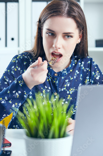 Business woman eating lunch at her workplace looking at the laptop screen. Folders with documents in the foreground. Dedline concept. photo