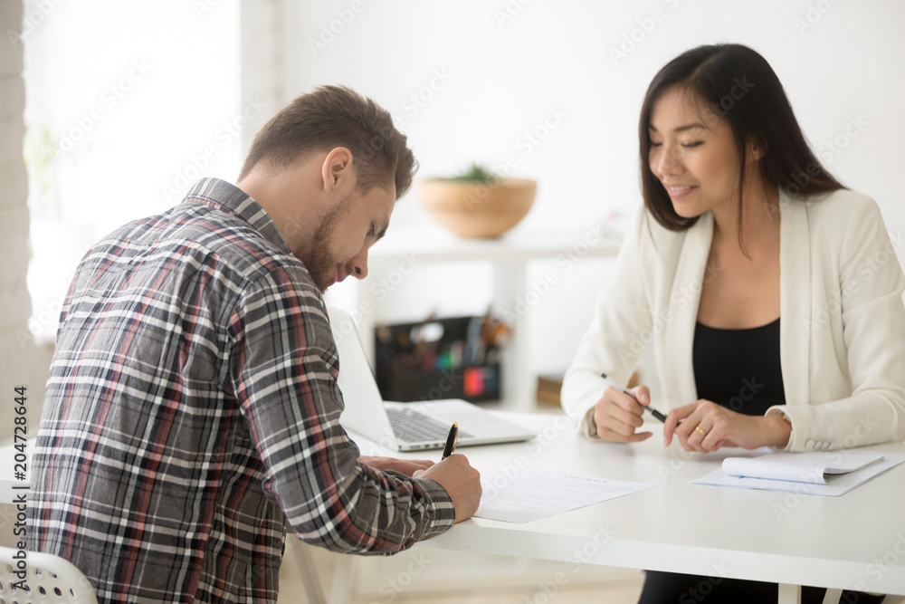 Caucasian businessman signing contract buying business insurance or taking loan at meeting with asian hr businesswoman, successful job candidate puts signature on employment agreement, hiring concept Caucasian businessman signing contract buying business insurance or taking loan at meeting with asian hr businesswoman, successful job candidate puts signature on employment agreement, hiring concept