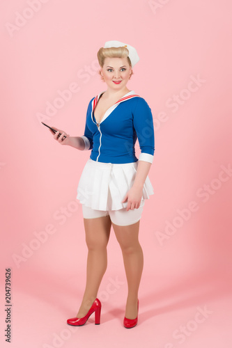 Fashion blonde dolly in sailor dress with smartphone and red heels isolated in studio on pink rose background photo