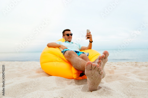 Cheerful young man takes a selfie, while listening to music in headphones. Sitting on yellow air sofa lamzac, on the sandy beach, by the sea. photo