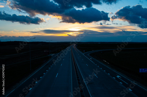 Asphalt road with four lanes leaving the sunset, blue sunset sky with the setting sun, hidden behind the clouds. Clouds illuminated by sunset light