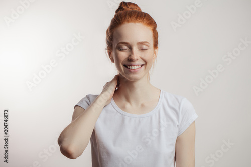 Pretty red-haired young woman with hair bun having joyful expression closing her eyes with pleasure and smiling dreamly isolated over white background photo