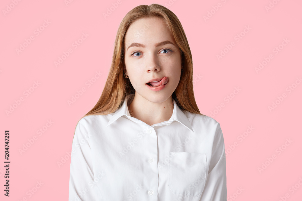 Lovely teenage girl licks lips, shows tongue, sees something pleasant or tasty, has appealing appearance, wears white shirt, isolated over pink background. Delighted female student poses indoor Lovely teenage girl licks lips, shows tongue, sees something pleasant or tasty, has appealing appearance, wears white shirt, isolated over pink background. Delighted female student poses indoor