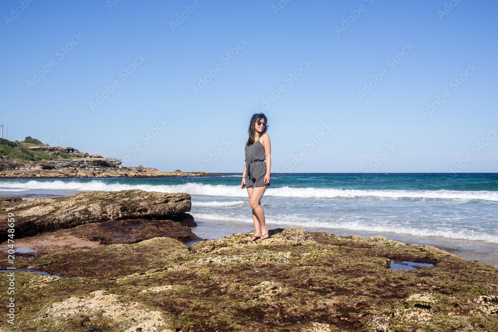 Young woman with sunglasses standing on the rocky shore of Maroubra beach, Sydney, Australia.