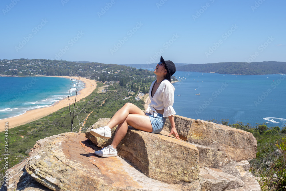 Young woman with sunglasses sits on the background of Palm beach on top of a rock at the lighthouse in Barrenjoey, Sydney, Australia. Young woman with sunglasses sits on the background of Palm beach on top of a rock at the lighthouse in Barrenjoey, Sydney, Australia.