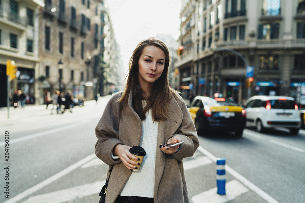 Fototapeta premium Young fashion blogger with long blonde hair is messaging to friend by a smartphone while crossing the road with a paper coffee cup in her hand. Attractive stylish girl is reading sms on mobile phone.