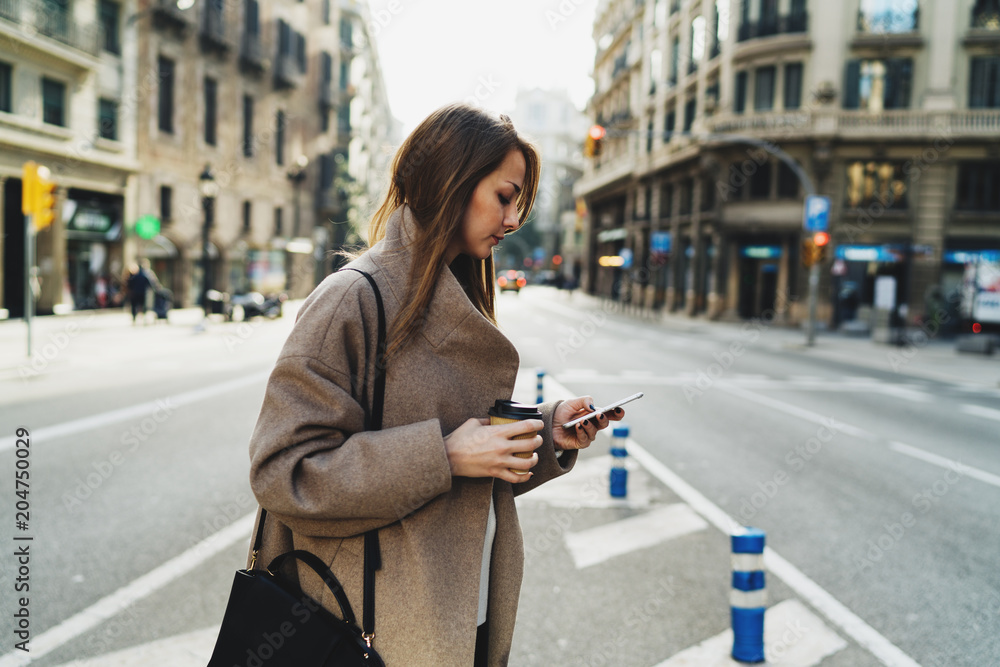 Model look woman reading emails on the smartphone while standing on a street background. Caucasian female texting messages on the mobile phone while crossing the street holding a take away coffee cup. Model look woman reading emails on the smartphone while standing on a street background. Caucasian female texting messages on the mobile phone while crossing the street holding a take away coffee cup.