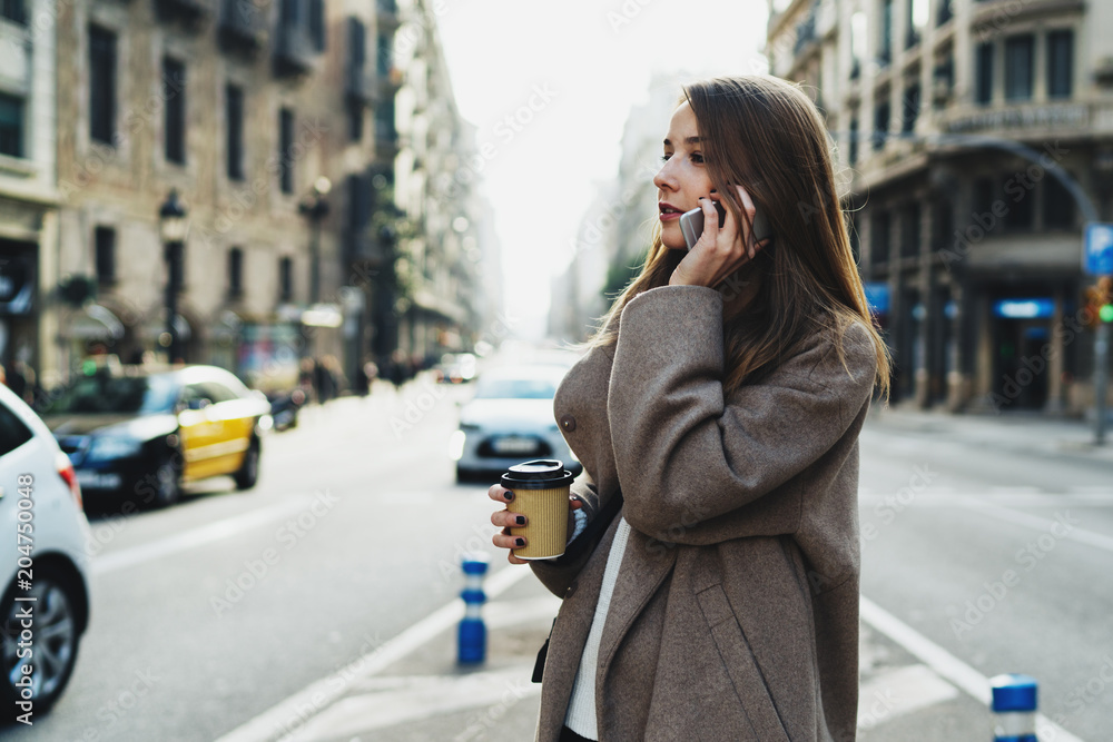 Obraz premium Beautiful caucasian female is talking by a smartphone while standing on the street with a take away coffee. Charming lady in stylish coat looking aside while chatting with a friend by a mobile phone.