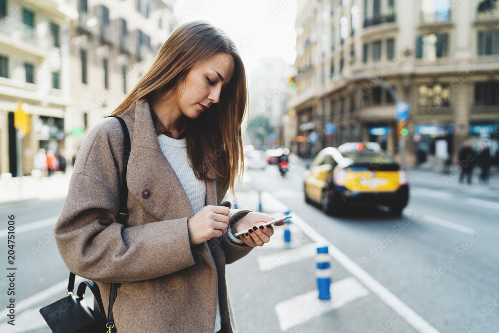 Obraz premium Successful designer reading email on mobile phone connected to wifi while walking the street. Charming student girl wearing stylish clothes searching information in the internet on a smartphone.