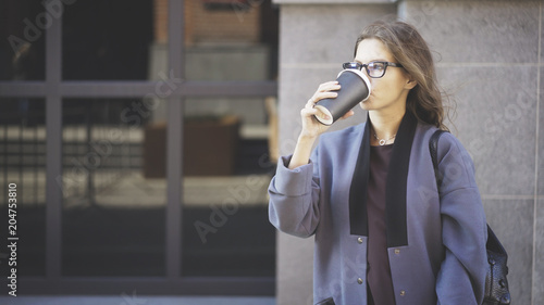 Businesswoman with long beautiful hair wearing glasses and a gray coat is drinking a cup of coffee outdoors. An autumn day.
