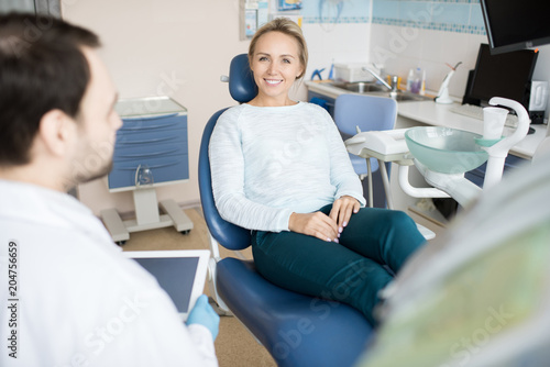 Beautiful woman sitting in chair of dentist smiling at camera while doctor using tablet in cabinet. 