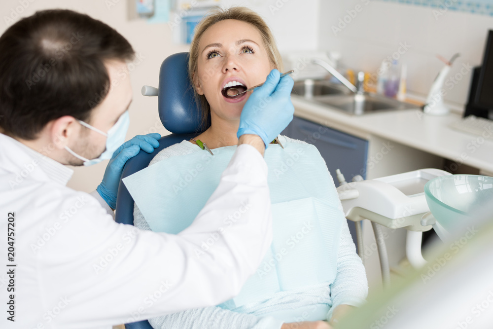 Confident man in gloves and mask using small mirror tool and exploring state of teeth of young woman on seat. Confident man in gloves and mask using small mirror tool and exploring state of teeth of young woman on seat.