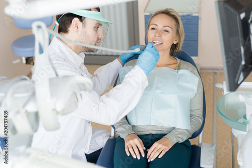 Professional dentist in uniform drilling tooth of pretty woman sitting in chair having visit. 