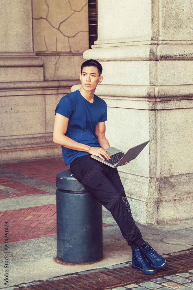 Way to Success. Young Asian American Man studying, working in New York, wearing blue T shirt, black pants, sneakers, sitting on pillar on street on campus, working on laptop computer, thinking..