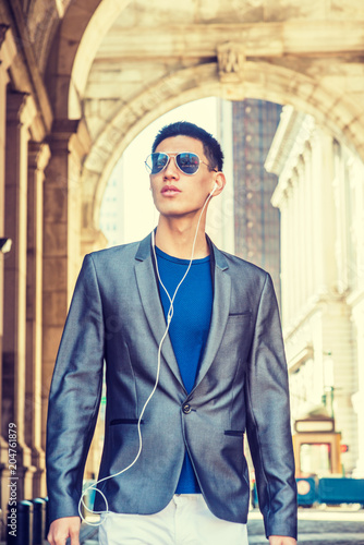 Young Asian American Man traveling in New York, dressing in gray blazer, white pants, wearing sunglasses, earphones, listening music, walking through small, narrow old street. Sunlight on background..