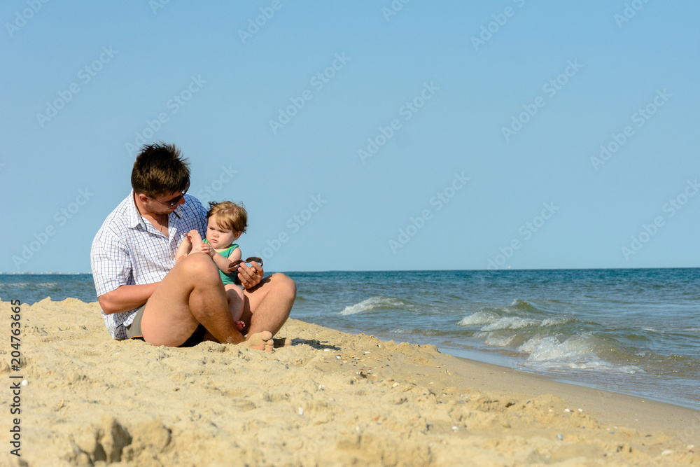 A father with a little daughter sits on the beach at the background of the sea A father with a little daughter sits on the beach at the background of the sea