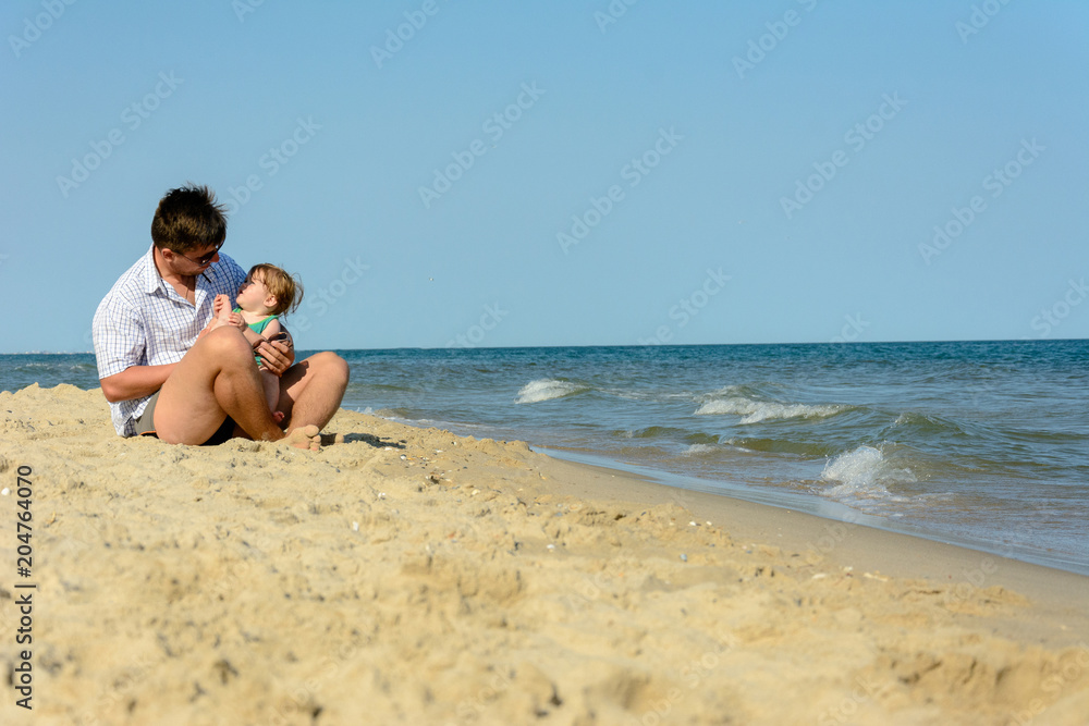 A father with a little daughter sits on the beach at the background of the sea
