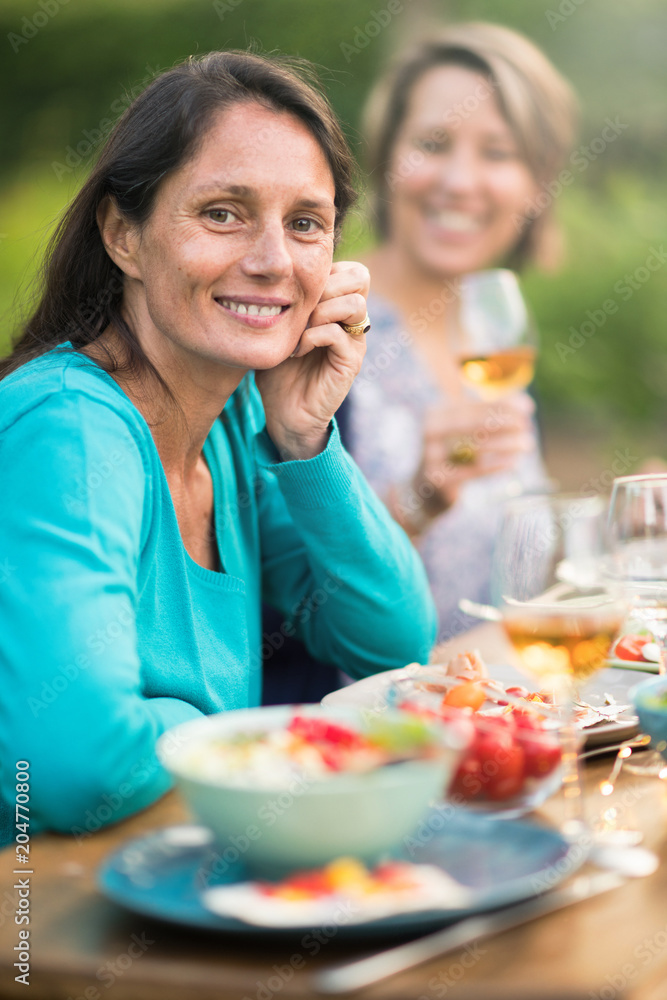 Looking at the camera. Portrait of a beautiful brunette woman in her forties. She sits around a table in a garden with friends for dinner. Looking at the camera. Portrait of a beautiful brunette woman in her forties. She sits around a table in a garden with friends for dinner.
