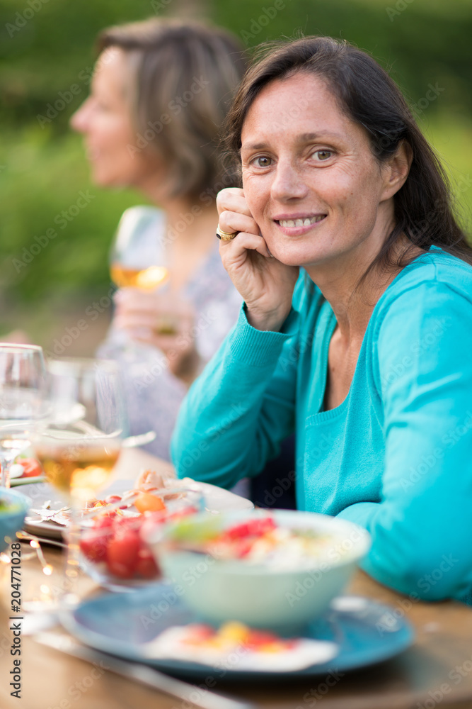 Looking at the camera. Portrait of a beautiful brunette woman in her forties. She sits around a table in a garden with friends for dinner.