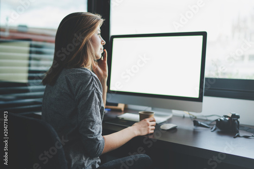view from back to female professional entrepreneur is talking on mobile phone sitting at desk in front of monitor with blank space for design. Mockup screen with copy space.woman makes a business call