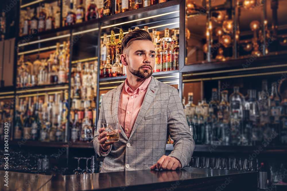 Stylish handsome male in an elegant suit holds a glass of whiskey at bar counter background.