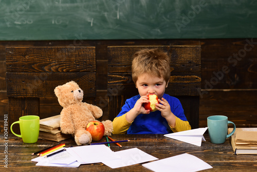 School break. Hungry kid biting apple in classroom. Small boy playing with paper plane and teddy bear photo