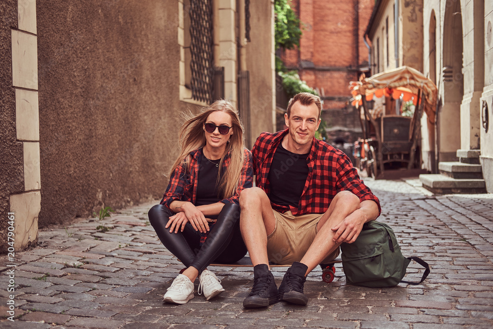 Young happy hipster couple, rest during a walk sitting on the pavement in the old Europe street.