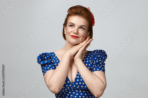 Horizontal shot of glamourous fashionable young European female wearing vintage dress with low cut and bright tmake up placing hands under her chin and looking up with thoughtful dreamy smile photo