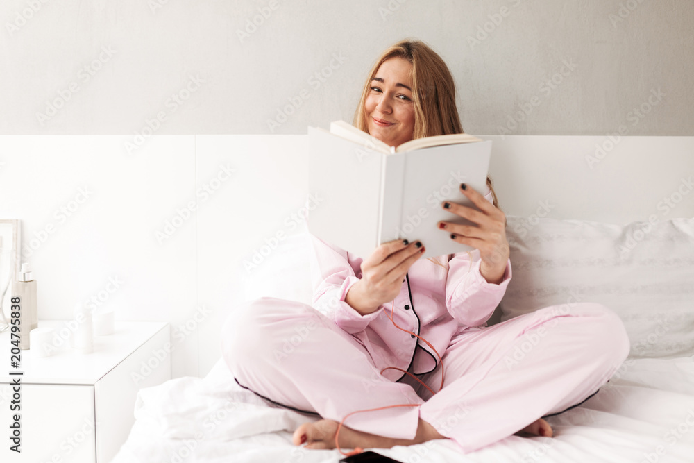 Portrait of young smiling woman sitting on bed and joyfully looking in book at home isolated Portrait of young smiling woman sitting on bed and joyfully looking in book at home isolated