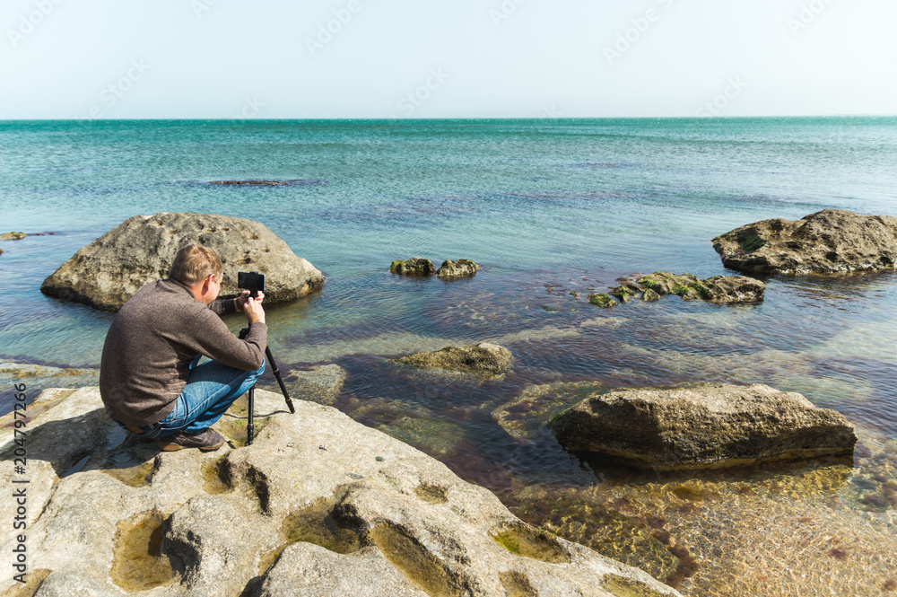 Man takes picture on the phone mounted on a tripod on a sea rocky coast