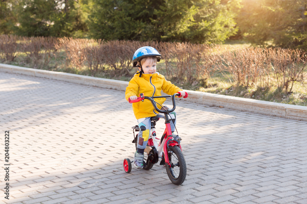 Three years old child boy in safety helmet riding on a bicycle at cobbles road in the city park in spring or summer. Happy childhood and kids first trip on a four-wheeled bicycle Three years old child boy in safety helmet riding on a bicycle at cobbles road in the city park in spring or summer. Happy childhood and kids first trip on a four-wheeled bicycle