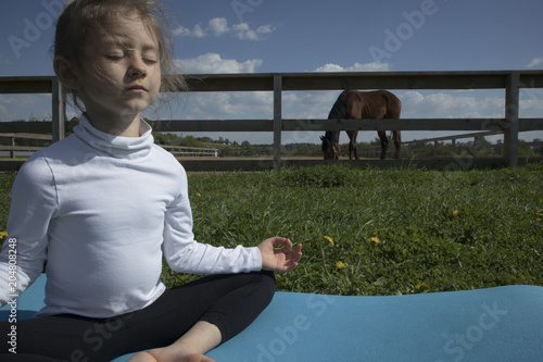 The young girl is engaged in gymnastics on the grass healthy lifestyle. Child girl doing gymnastics. Little cute girl practicing yoga pose on the background of the paddock with a horse photo