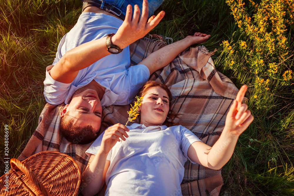 Fototapeta premium Young couple relaxing after having picnic. Woman and man lying on grass and talking at sunset. Guys pointing at sky