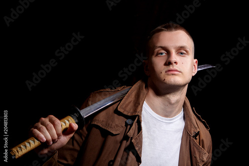 Young man dressed in a brown coat and white t-shiert, holding a katana.Modern samuria.Fashion portrait.Urban warior.Dramatic light photo