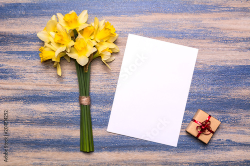Fototapeta Naklejka Na Ścianę i Meble -  Workplace with a gift with a red ribbon, a piece of paper and a bouquet of daffodils on a blue wooden background. Flat lay design, top view.