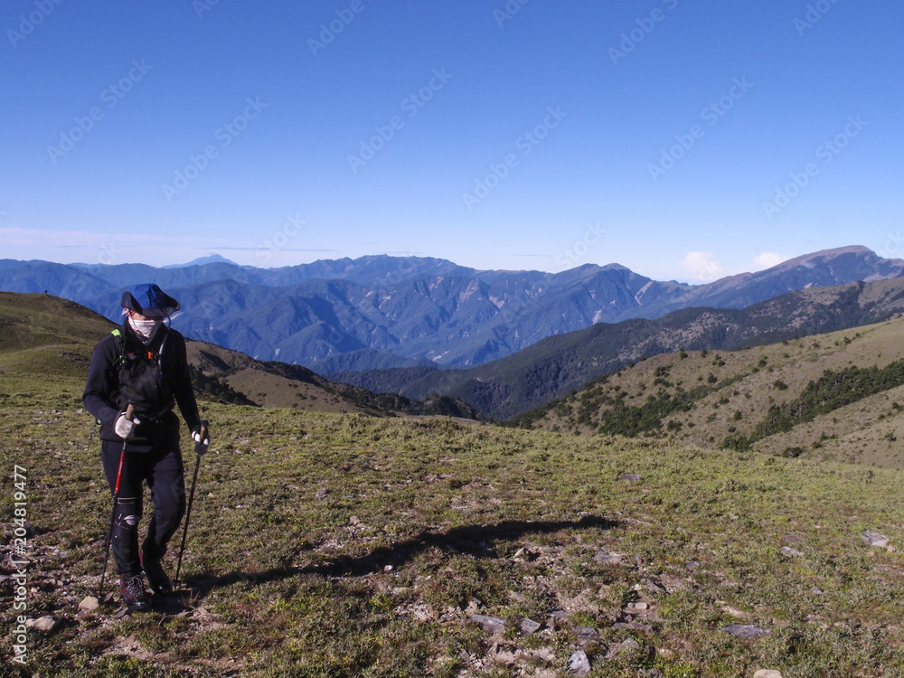 A male hiker in a hat holding trekking poles and walking on a trail. Background is the beautiful alpine landscape of Taiwan with a clear blue sky.