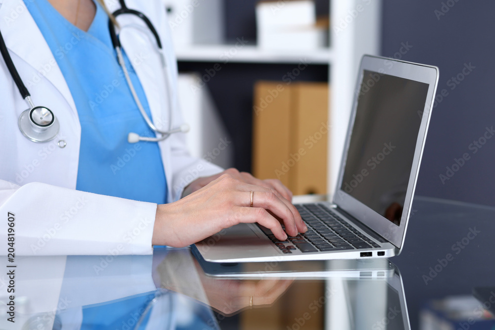 Doctor typing on laptop computer while sitting at the glass desk in hospital office. Physician at work. Medicine and healthcare concept Doctor typing on laptop computer while sitting at the glass desk in hospital office. Physician at work. Medicine and healthcare concept