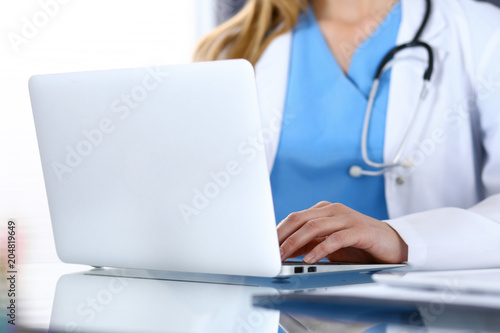 Doctor typing on laptop computer while sitting at the glass desk in hospital office. Physician at work. Medicine and healthcare concept