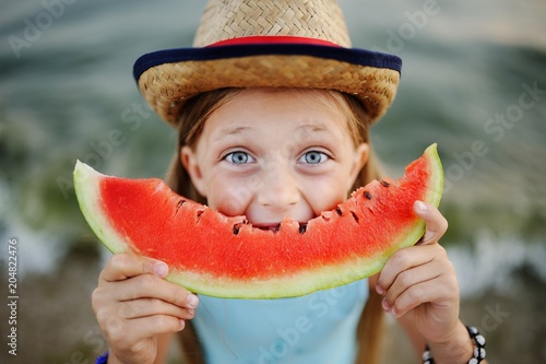 baby girl in the hat eagerly eats juicy watermelon on sea background
