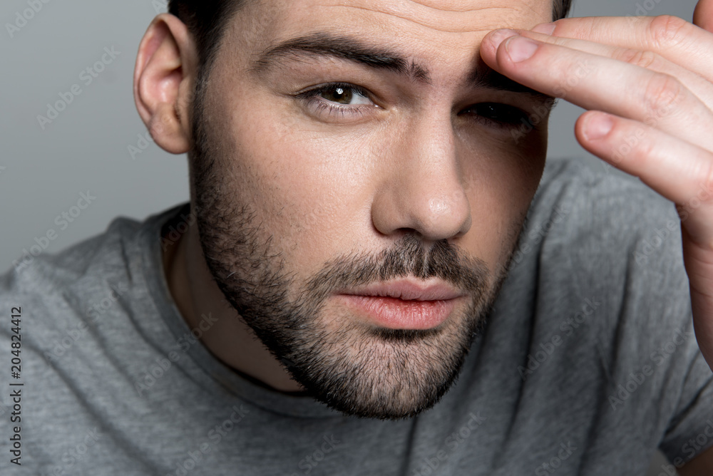 Obraz premium Ache concept. Close up portrait of young exhausted man is looking at camera while suffering from headache. He is touching his forehead. Focus on his face