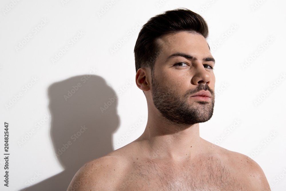 Obraz premium Portrait of stylish young bearded man is standing against white wall and looking at camera seriously. Copy space in the left side. Beauty concept