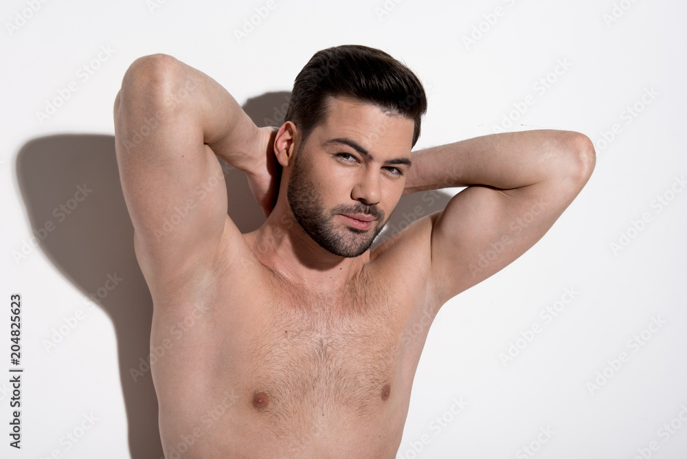 Perfect skin. Portrait of charming topless guy is standing against light background and demonstrating his smooth armpits. He is raising his arms up while looking at camera confidently Perfect skin. Portrait of charming topless guy is standing against light background and demonstrating his smooth armpits. He is raising his arms up while looking at camera confidently