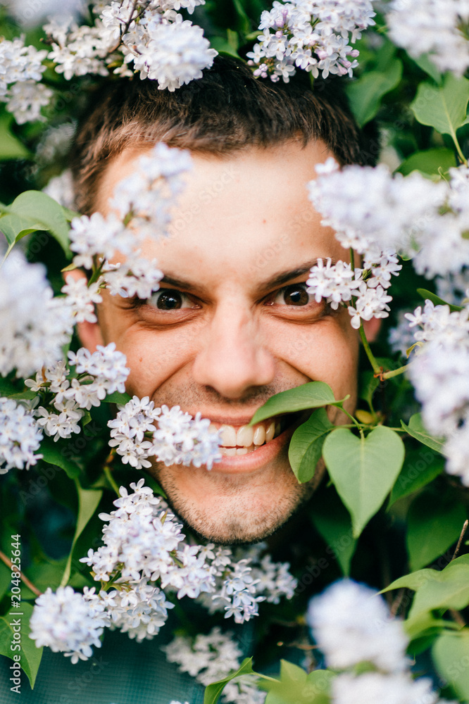 Obraz premium Closeup odd unusual male portrait outdoor at nature in beautiful lilac bushes. Adult funny man face grimace surrounded by blooming flowers. Emotions and facial expxression. Clowning and fooling.
