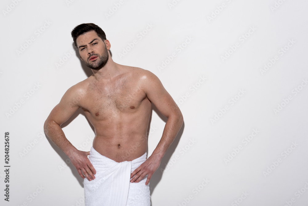 Beauty concept. Portrait of young topless man is standing with towel against light background. He is looking at camera seriously while holding hands on hips. Copy space in the right side Beauty concept. Portrait of young topless man is standing with towel against light background. He is looking at camera seriously while holding hands on hips. Copy space in the right side