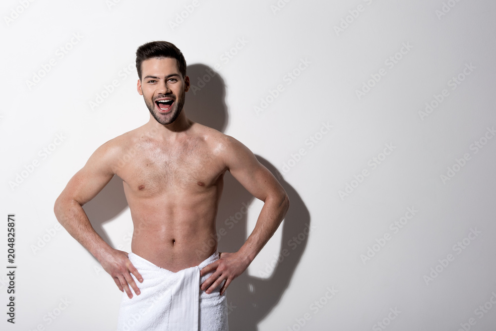 Full of joy. Portrait of happy cute bristled topless guy is standing in towel and looking at camera with wide smile. He is holding hands akimbo while demonstrating his muscular body. Copy space