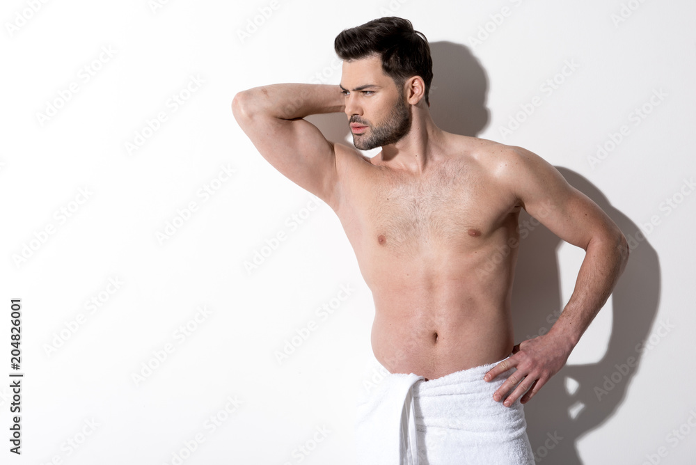 Perfection. Gorgeous cute guy with stubble is standing with towel on his hips and looking aside dreamily. He is holding hand behind his head. Copy space in the left side Perfection. Gorgeous cute guy with stubble is standing with towel on his hips and looking aside dreamily. He is holding hand behind his head. Copy space in the left side