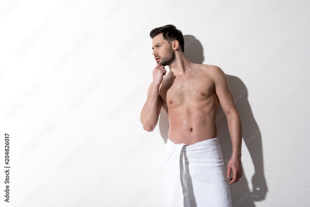 Fototapeta premium What is this. Young charming guy is standing with towel on hips and looking aside with concentration. He is touching his chin while posing against light background. Copy space in the left side