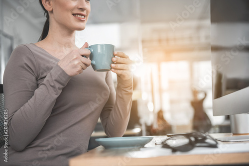 Happy female drinking mug of appetizing beverage while looking at digital device in office. Satisfied employer at labor concept